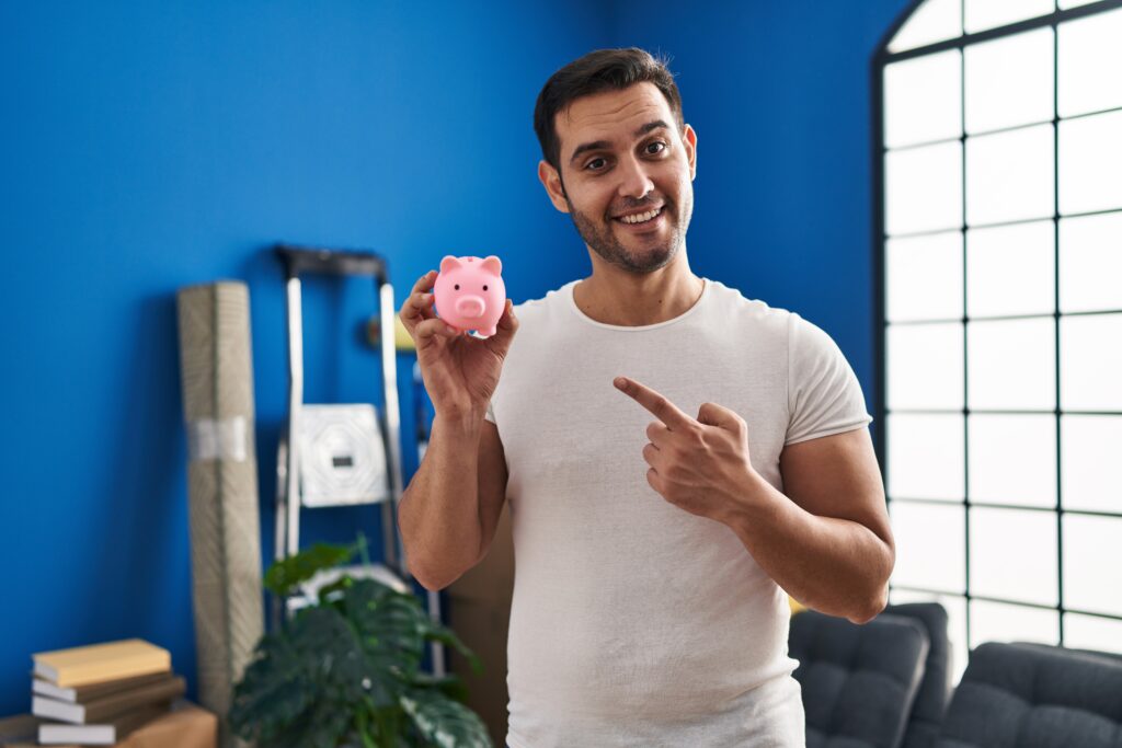 Young hispanic man with beard holding piggy bank at new home smiling happy pointing with hand and finger